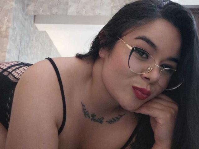 EveWillis with glasses, pouty red lips, black fishnet top, tattoos above her tits, leaning on her hand, brown hair, light skin, sexy, looking at the camera.