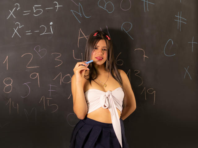 SheylaSpooner with long brown hair, red bow, white strapless top, blue skirt, holding a ruler, smiling at a chalkboard with math equations. She has cute tits and a sexy face.