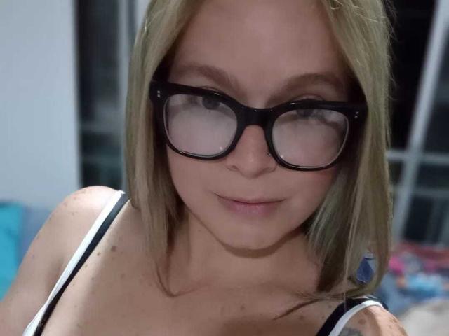 PamelaColin with glasses, brown hair, slightly flushed face, looking up, small nose, subtle smile, wearing earrings, soft light, blurry background, upper body, casual, slight pinkish skin tone.