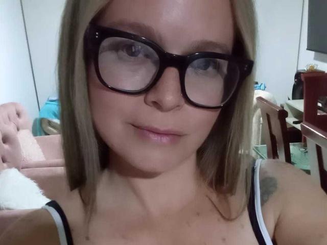 PamelaColin has brown hair, large black glasses, and a white tank top showing her small tits. She's looking down with a slight smile. Her skin is light.