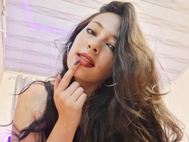 CoquineElise de rodillas, con una falda de red rosa que deja ver su trasero, sobre una cama, con una manta blanca, un teclado negro y una puerta en el fondo, con su cabello rubio y piel clara..