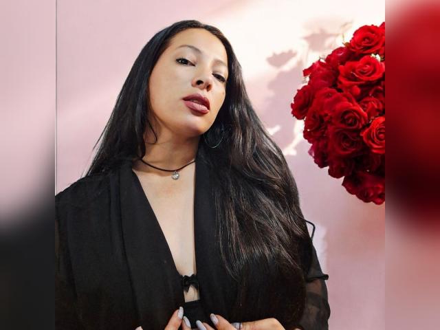 GiaGold with long brown hair, red lipstick, and hoop earrings, touching her lip with a finger, looking at the camera with a slightly winking, sexy expression.