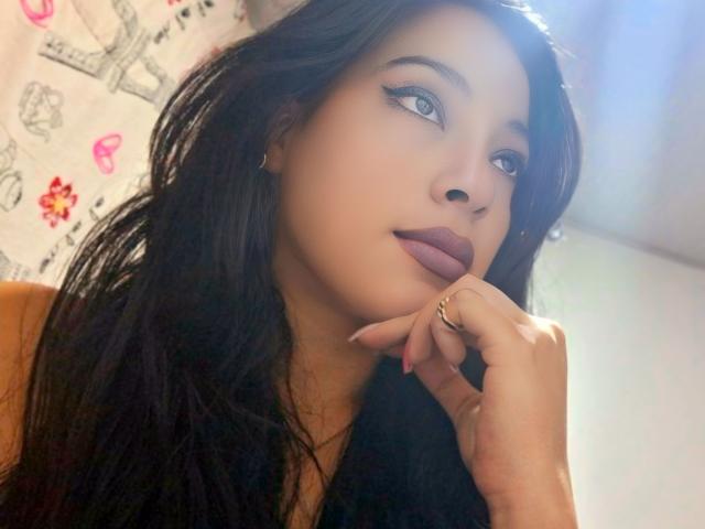 GiaGold tiene el cabello oscuro y ondulado, lápiz labial rojo y labios rosados ligeramente separados. Está mirando a la cámara con una expresión relajada, casi juguetona. Su mano derecha está cerca de su rostro..