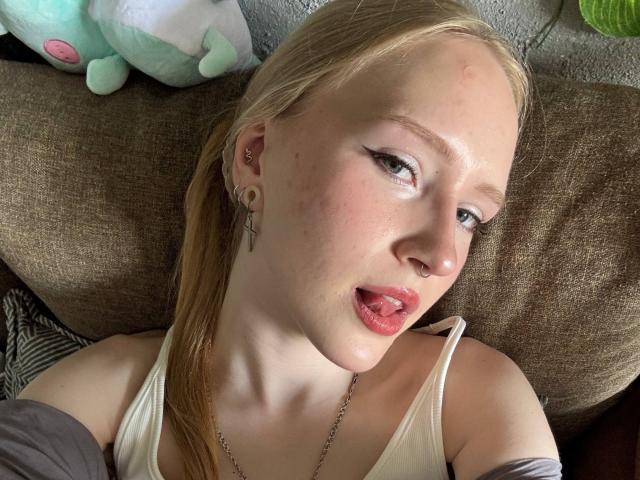 SiennaHays with long brown hair, wearing a sparkly, mesh, halter top that exposes her tits, black shorts, and a tattoo on her right arm, sitting and looking at camera.