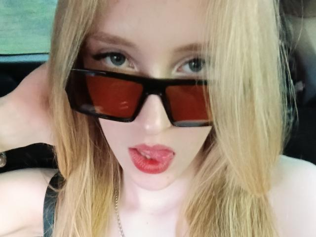 NievesLamanna has blonde hair, wearing black glasses with red lenses, red lipstick, and a black strap top, with her hand in her hair, looking at the camera.