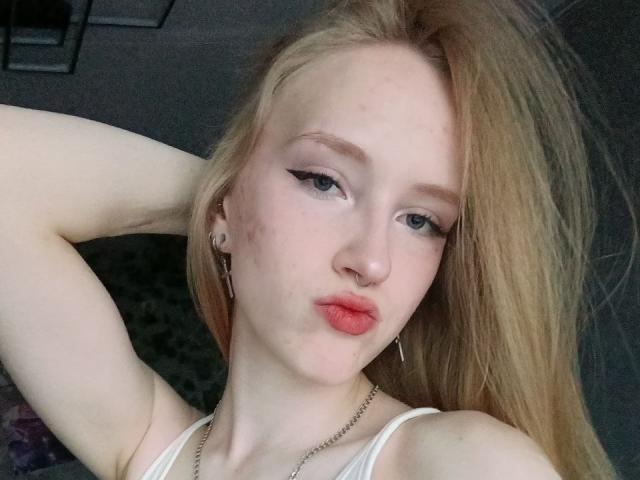 NievesLamanna with pale skin, blonde hair, and pink lipstick, pouting with arms behind head, wearing white tank top, silver earrings, and chain necklace, looking at camera.