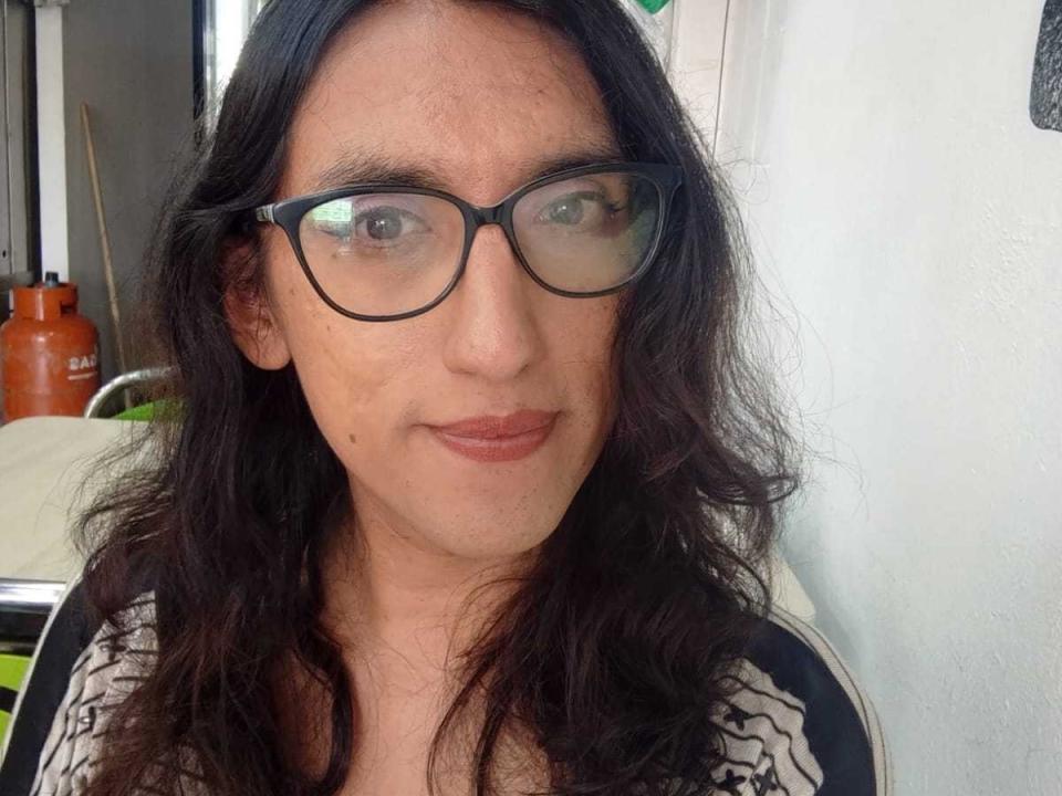 AlejaSexy69 tem cabelos castanho-escuros ondulados, usa óculos pretos e tem um sorriso discreto com batom rosa claro. Seus seios são pequenos e ela está vestindo uma blusa preta e branca..