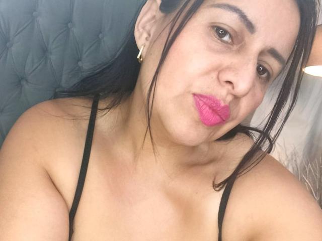 IvonnaLune indossa biancheria intima di pizzo nero, mostrando il suo seno prosperoso, le labbra rosa e i capelli scuri, mentre è seduta su una poltrona grigia imbottita. La sua pelle è chiara e indossa orecchini dorati..