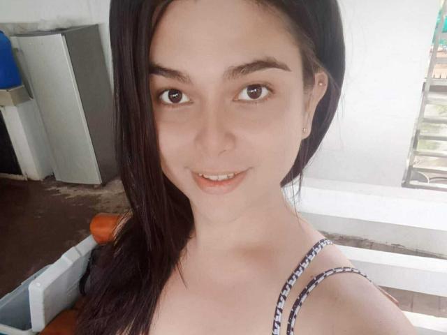 TsRosanna, com pele clara, olhos castanhos e cabelos longos e escuros, sorrindo para a câmera, vestindo uma blusa listrada, usando pequenos brincos e com uma expressão adorável e relaxada..