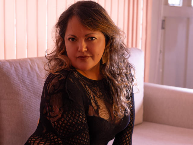 LaurenPascal, avec ses cheveux bruns ondulés, portant une robe noire en résille, laissant entrevoir son soutien-gorge noir et exposant ses fesses, assise sur un canapé beige, regardant la caméra..
