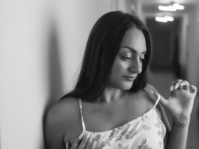 Schwarz-weißes Foto von HannahRosee mit langen, dunklen Haaren, die nach unten schaut, in einem blumigen Spaghetti-Trägerkleid, eine Zigarette in der Nähe ihres Schulters haltend, ein leichtes Lächeln, weiches Licht, Tätowierung am Handgelenk..