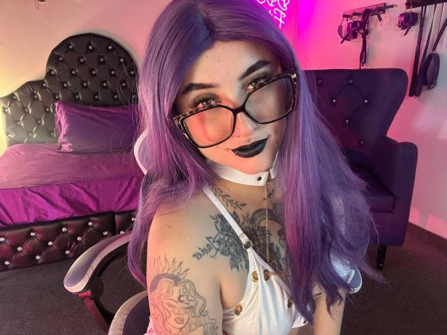 LilithDark69 met paars haar, een bril, zwarte lippenstift, een witte leren riem en een korte broek, zittend op een stoel, met tatoeages zichtbaar, in een kamer met neonverlichting en donkere meubels..