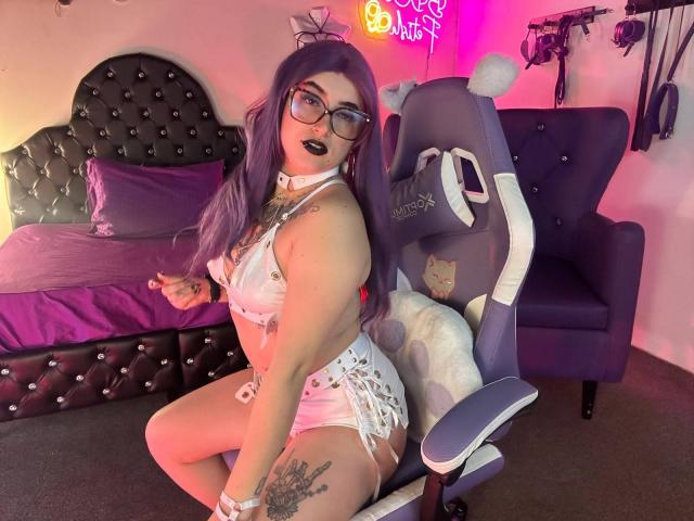 LilithDark69 met paars haar, zwarte lipstick, een bril en tatoeages, draagt een witte jurk en zit op een stoel, waarbij ze haar tatoeages op haar borsten en billen laat zien, in een kamer met roze verlichting..