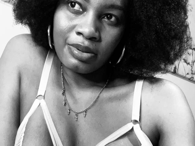 MelaninDose, avec des cheveux crépus et afro, porte un soutien-gorge blanc à bretelles fines, des boucles d'oreilles cerceaux et un collier avec de petites croix, et a une expression légèrement boudeuse. Ses seins sont visibles à travers le soutien-gorge..