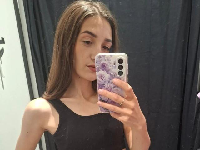 JoannaVanderheide haciendo sentadillas en un gimnasio, vestida con un top deportivo y pantalones cortos negros, zapatillas blancas, sosteniendo un teléfono, con cabello castaño largo y una expresión concentrada..