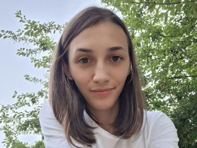 Joanna Vanderheide, con lunghi capelli castani, indossa una camicia a righe blu, pantaloni bianchi e tiene in mano una borsa verde, mentre si scatta un selfie in un ascensore di metallo, sorridendo..