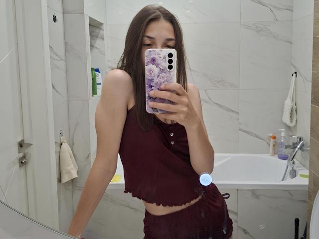 Het gezicht van JoannaVanderheide, met halflang bruin haar, gekleed in een witte blouse, met kleine oorringen en een subtiele glimlach, op de achtergrond van groene bladeren..