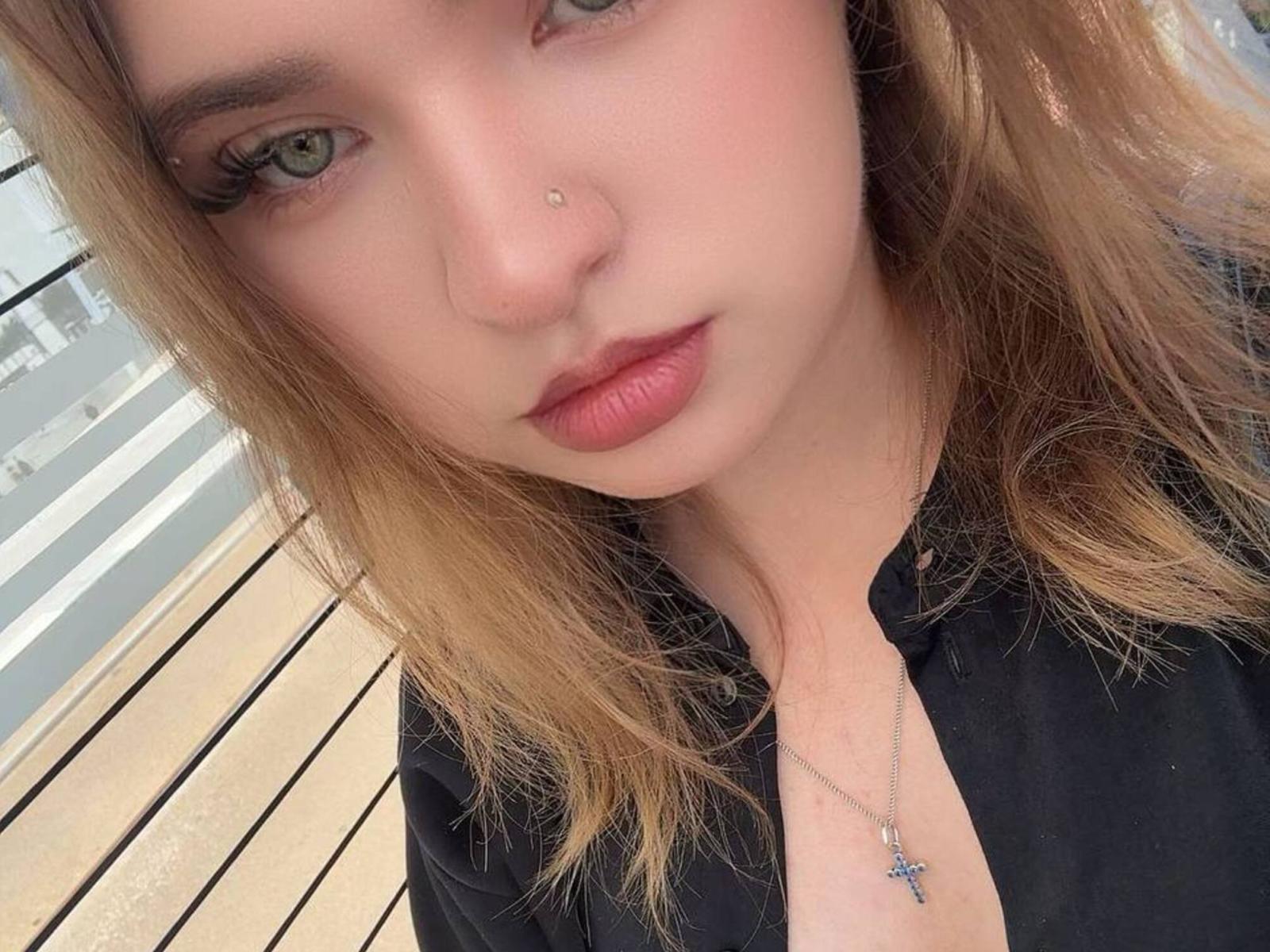 Susana Pizani tem um piercing no nariz, olhos verde claro e lábios rosados. Ela está vestindo uma camisa preta com um colar de cruz prateado, e seu cabelo loiro é levemente ondulado..