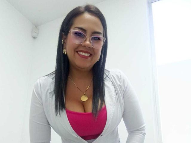 Kathiusca with medium tits, wearing a pink top, white cardigan, glasses, gold earrings, and necklace, smiling in a white room.