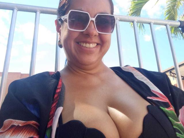Kathiusca, com seios grandes, sorrindo, usando óculos de sol brancos, blusa preta com estampas vermelhas e verdes, e brincos pretos, em pé ao lado de um corrimão branco, céu azul ensolarado, folha de palmeira visível..