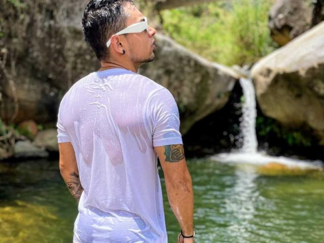 Xanderrt draagt een nat, doorschijnend wit T-shirt, waardoor zijn strakke billen en tatoeages zichtbaar zijn, met een waterval op de achtergrond. Hij kijkt om, en draagt witte zonnebrillen..