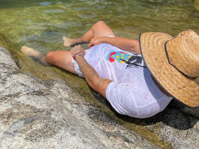 Xanderrt's wet white shirt clings to their chubby ass and tattooed arms, wearing a straw hat, lying in shallow green water with legs spread, on a rocky surface.