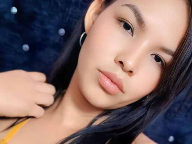 Delilath's face with light brown skin, straight black hair, small nose, full pink lips, brown eyes, wearing a yellow top, black ear ring, hand on neck.