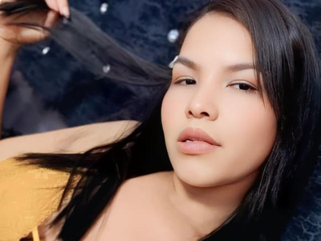 Delilath has light brown skin, black straight hair, and is holding a lock of it with her right hand. She's wearing a yellow top, with slightly parted pink lips.