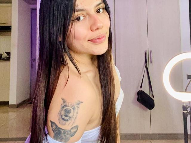 SophiaMarx with long black hair, light skin, small nose, and a tattoo of a dog's face on her upper arm, smiling at the camera, wearing a white off-shoulder top.