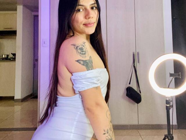 SophiaMarx with long black hair, white off-shoulder dress, tattoos on shoulder and arm, smiling at camera, ring light on right, standing in room. Boobs visible from side.