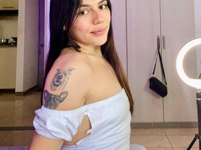 SophiaMarx with long black hair, light blue off-shoulder top, white pants, tattoos on arm and shoulder, smiling at ring light, showing her ass, in a modern room.