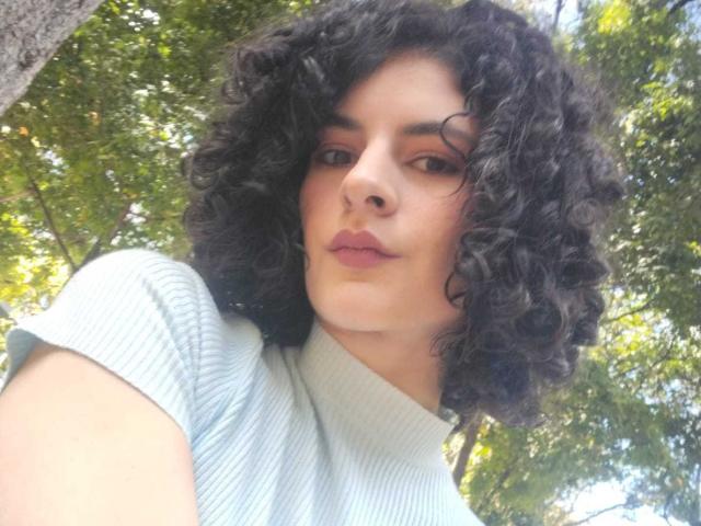 RoxyGraff with curly black hair, light skin, wearing a ribbed light green turtleneck, has full lips and a slight smile, looking down at the camera.