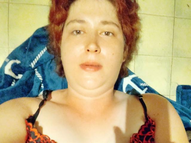 Lucy-2026-hot with red hair, pale skin, wearing a floral tank top, sitting on a black leather couch, smiling slightly, showing small tits, and a black earbud.