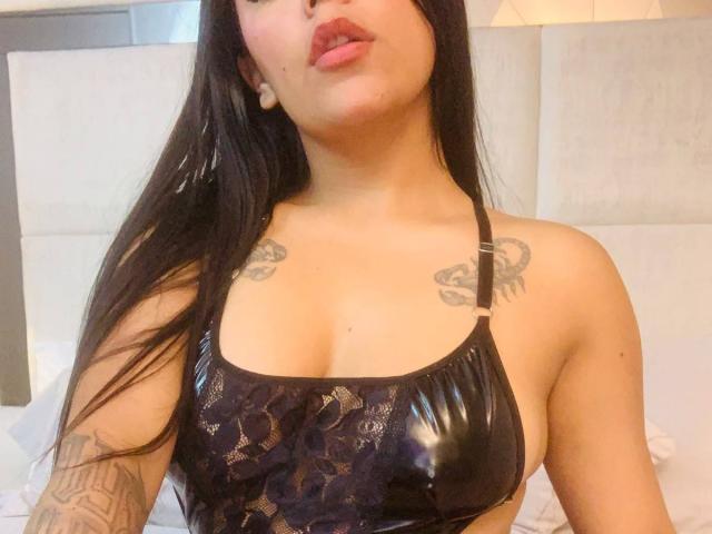 Topless SweetClaraa with curly brown hair, sticking out tongue, medium tits, light skin, small nipples, shadowed chest, and a slight smile, in a dimly lit room.