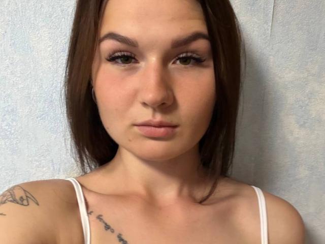 LindsySwisher has brown hair, green eyes, and a neutral expression. She's wearing a white top with tattoos on her right shoulder and upper chest. Her tits are small.