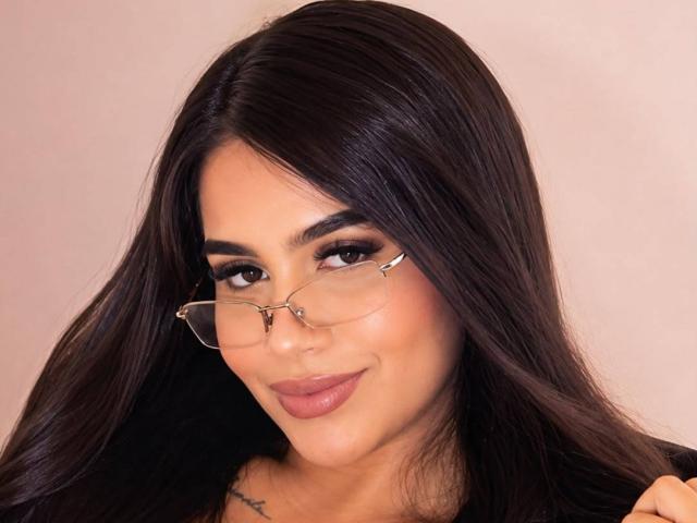 Isis Ferrer, met donker, glanzend haar, die een bril draagt, heeft een smoky oogmake-up, volle, roze lippen en een tatoeage op haar sleutelbeen, en ze glimlacht subtiel..