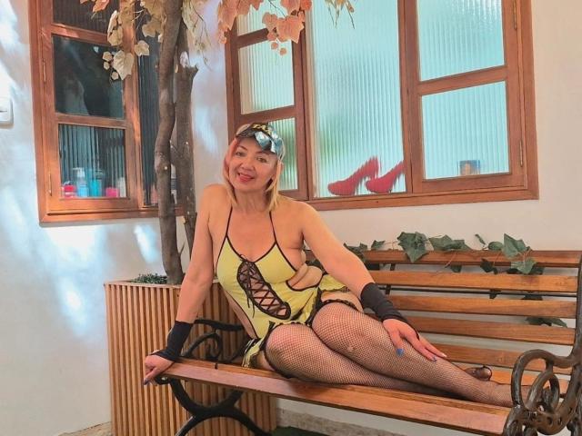AmelieLux with blonde hair, wearing a yellow, cut-out, lace-up dress, black wristbands, and fishnet stockings, sitting on a wooden bench, smiling, with her legs crossed.