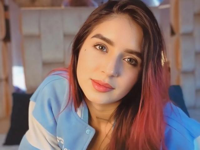 LeoFire with red-tipped brown hair, wearing a blue and white shirt, has pink lips, brown eyes, and a small mole on her right cheek. She's smiling softly.