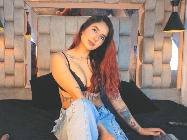 LeoFire with red and black hair, wearing a black bra, showing her tits, tattoos on arms and side, sitting on a black bed, blue jeans, looking at camera.