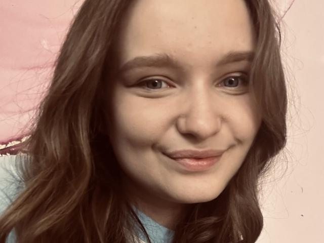 MagikPeachh hat hellbraunes, welliges Haar, ein leichtes Lächeln und braune Augen. Sie trägt ein blaues Hemd mit einem rosa Hintergrund. Ihre Nase ist klein und sie ist süß..