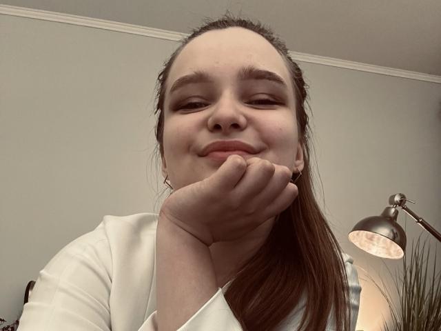 MagikPeachh with hand under chin, smiling slightly, brown hair in ponytail, white shirt, soft light from lamp on right. She has fair skin and small, natural tits.