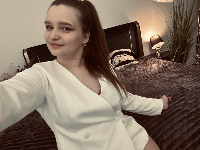 MagikPeachh con una lunga coda di cavallo marrone, giacca bianca con bottoni, reggiseno rosso che spunta, seduta sul letto, sorridente, con un braccio teso per un selfie, coperta trapuntata grigio scuro sullo sfondo..