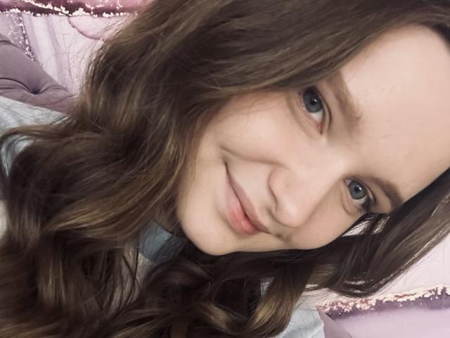 MagikPeachh has long brown wavy hair, light skin, blue eyes, and a subtle smile. She's wearing a light blue shirt with a purple lace trim.