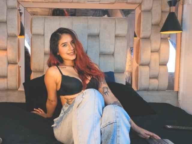 LeoFire, con i capelli rossi, un reggiseno nero e jeans blu, è seduta su un letto, sorridendo, mostrando tatuaggi, piccoli seni e un bel sedere, con un cuscino nero e una lampada..