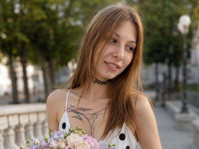 JaPorada with light brown hair, smiling, wearing a white dress with black dots, choker, and holding a flower bouquet, showing tattoos on her chest, in a park.