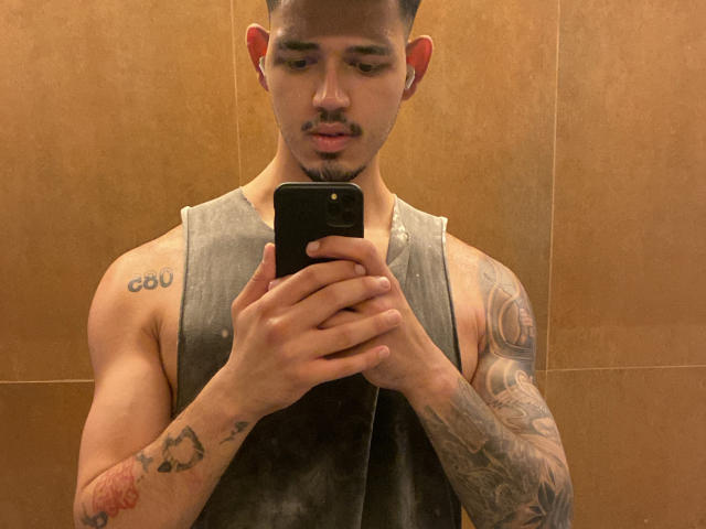 RodrigoHot69 macht ein Selfie im Spiegel, trägt ein schmutziges, graues Tanktop, zeigt Tattoos auf beiden Armen, hält ein schwarzes Telefon, hat braune Haut, kurze Haare und einen Bart..