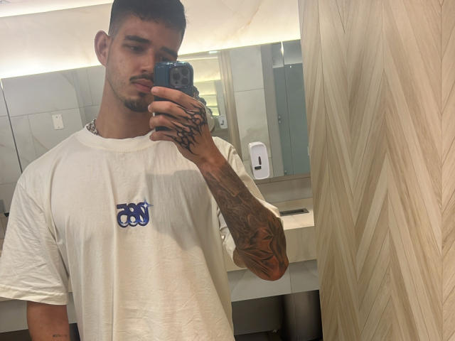 RodrigoHot69 with tattoos on arm, white tee, and short dark hair, taking mirror selfie with phone in modern bathroom, wood-patterned wall on right.