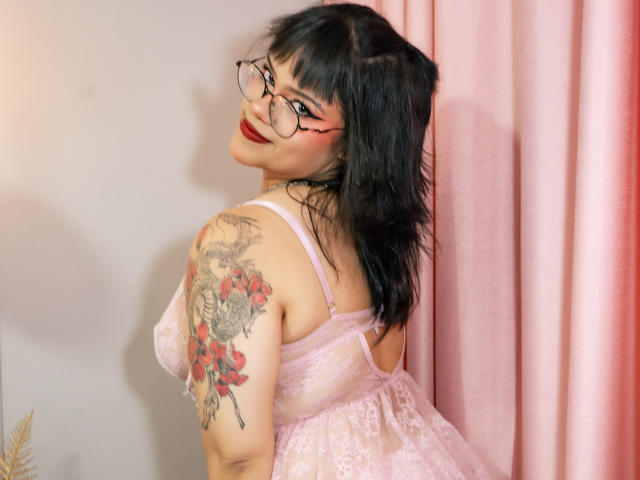 AnneStonee with black hair, glasses, red lipstick, pink lace lingerie, and a large floral tattoo on her right arm, smiling over her shoulder, showing her tits.