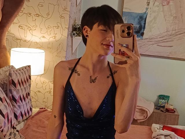 LissyLuxe with short dark hair, wearing a deep blue sparkly dress, showing a tattooed chest and tongue out in a mirror selfie. She has small breasts and is holding a phone.