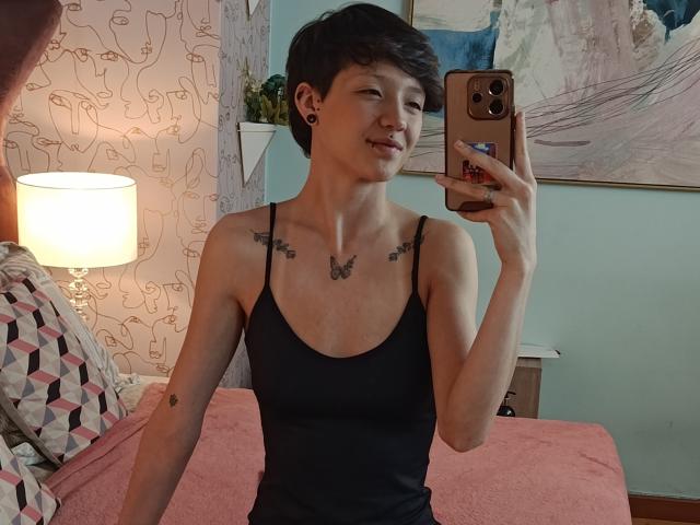 LissyLuxe com cabelo preto curto, seios pequenos, top de tanque preto, tatuagens de borboleta, segurando um telefone, sorrindo, cama com cobertor rosa, luminária, papel de parede estampado e pintura abstrata ao fundo..